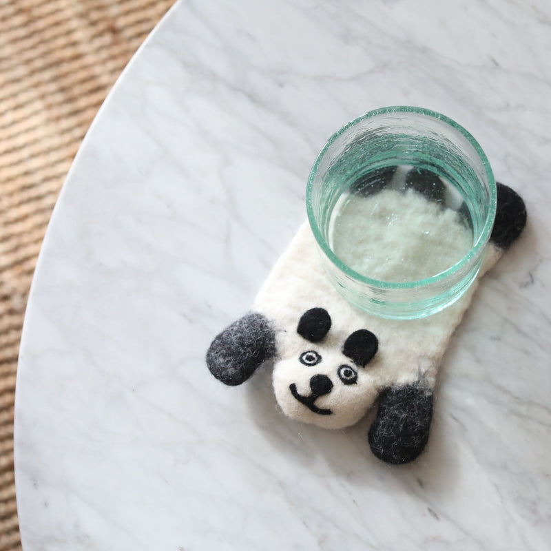 One unique, handcrafted felt panda coaster rests on a white marble table, holding a clear glass filled with water. This whimsical wool felt coaster, ethically handmade, features a predominantly white panda design with contrasting black ears, black eyes, and a charming, needle-felted smiling face.