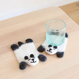 Two handmade wool felt panda coasters rest on a light wooden surface. One panda coaster is beneath a clear glass of water, showcasing its functionality. Each coaster features a simplified white panda body with black ears and paws, and needle felted facial details. These charming, ethically handcrafted coasters add a cozy, minimalist touch.