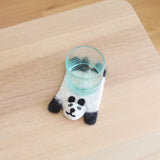 A whimsical felt panda coaster sits on a light wooden table, placed beneath a clear glass. This handmade wool felt coaster features a white panda body with black limbs and eye accents, detailed with a small, endearing smile. Crafted from sheep wool by Nepalese female artisans, it adds a charming touch.