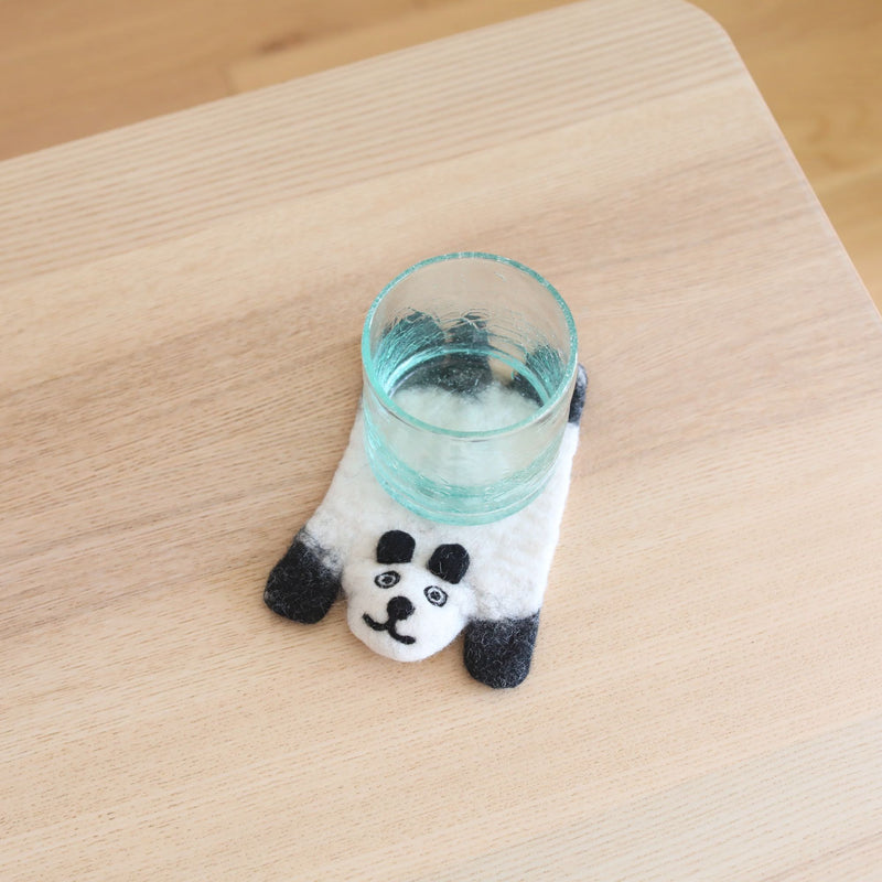 A whimsical felt panda coaster sits on a light wooden table, placed beneath a clear glass. This handmade wool felt coaster features a white panda body with black limbs and eye accents, detailed with a small, endearing smile. Crafted from sheep wool by Nepalese female artisans, it adds a charming touch.