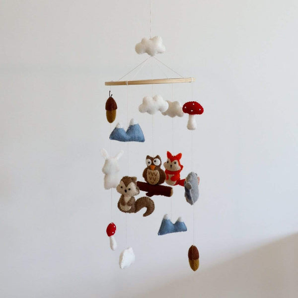 A Felt Woodland Animal Baby Mobile hangs from a white wooden hook, featuring a fox, owl, rabbit, and deer crafted with attention to detail using 100% New Zealand wool felt. The mobile is suspended in a minimalist setting with a plain white wall providing a neutral backdrop for the colorful and textured animals.