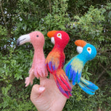 A needle felted flamingo finger puppet is held by a female hand, presented against a lush green foliage background. This vibrant pink flamingo puppet features a long neck and a distinctive white beak, showcasing its unique handcrafted details. Displayed on the fingertips, this soft, artisanal felt toy for kids is perfect for imaginative play.