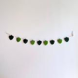 A greenish felt Monstera leaf garland hangs against a plain white wall, showcasing a natural, simple aesthetic. It features alternating felt leaves in two shades of green, connected by a thin brown jute string. A clear adhesive hook supports the garland.