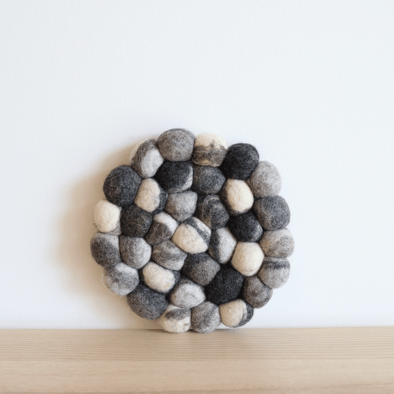 A thick felt ball pebble-stone pot trivet rests on a light wood surface against a minimalist white wall, creating a calm, natural aesthetic. This round, handcrafted wool felt pot trivet features a dense arrangement of organic wool felt balls in varying shades of gray, white, and black, giving it a textured, stone-like appearance. Ethically handmade by Nepalese female artisans using eco-friendly dyes, this unique trivet adds organic warmth to the setting.